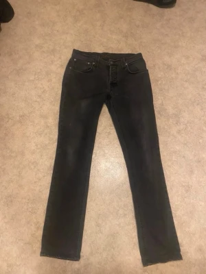 Svarta raka jeans från Nudie Jeans - Säljer ett par svarta jeans från Nudie Jeans med klassisk femficksdesign och raka ben. Jeansen har diskreta slitningar och är tillverkade i mjuk denim. Perfekta för dig som gillar en stilren och enkel look.