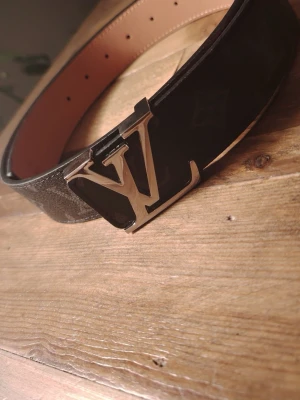 Louis Vuitto  - Snyggt svart skärp från Louis Vuitto med ikoniskt Lspänne i metall. Bältet har en klassisk design med brunt innerfoder och diskret monogrammönster på utsidan. Perfekt accessoar för att lyfta din outfit.