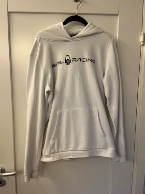 Vit hoodie från Sail Racing - Vit hoodie från Sail Racing med svart logotyptryck på bröstet och en mindre logga på huvan. Tröjan har en klassisk känguruficka framtill. Den har använts en del och har en liten fläck på axlen annars är den i bra skick. Hör av er om ni har frågor! 