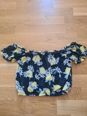 Blommig offshoulder topp med citroner - Söt offshoulder topp med puffärmar och elastiska kanter. Svart botten med gult citron- och blommönster. Perfekt för sommarens alla tillfällen och ger en fräsch vibe till din outfit.