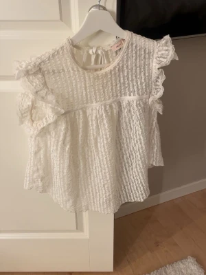 Vit spetsblus med volangärmar - Supersöt vit blus i spets med volangdetaljer på ärmarna och knytning i nacken. Blusen har en luftig och lös passform, rund halsringning och romantisk känsla. Perfekt att styla med jeans eller kjol för en trendig look.