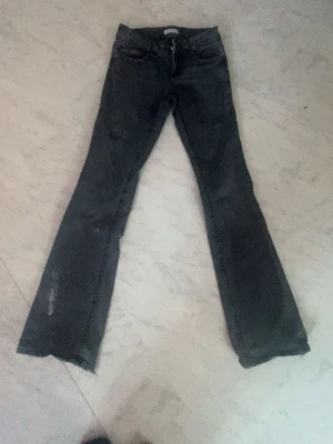 Grå bootcut jeans med fickdetaljer - Säljer ett par grå bootcut jeans med snygga fickor bak som har lock och knapp. Jeansen har en klassisk femficksmodell och är tillverkade i denim med lätt tvättad look. Perfekta för dig som gillar en avslappnad men trendig stil.