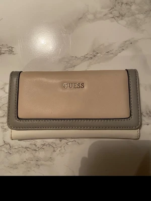 Beige och grå plånbok från Guess - Snygg plånbok från Guess i beige och grått konstläder med silvrig logga framtill. Plånboken har ett stilrent, rektangulärt format och dragkedjefack på baksidan. Perfekt för att hålla ordning på kort och kontanter.