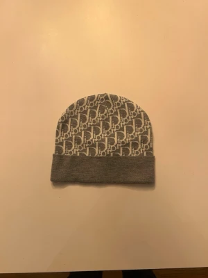 Grå Dior-mössa med logomönster - Snygg grå mössa från Dior med ikoniskt vitt logomönster över hela ytan och vikt kant. Tillverkad i mjukt material som känns bekvämt på huvudet. Perfekt accessoar för dig som vill ha en stilren och trendig look.