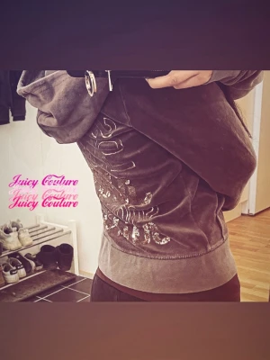  Vintage Juicy Couture grå hoodie  - Mjuk och lyxig vintage grå hoodie från Juicy Couture i velour med stor huva. Baksidan är dekorerad med glittrande guldpaljetter och text som ger en glamorös vibe. Perfekt för dig som gillar statement-plagg och vill sticka ut med en ikonisk look. 