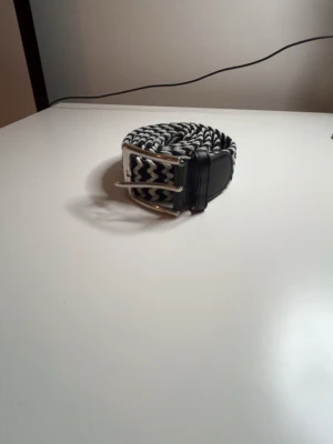 Svartvitt flätat bälte med zigzag-mönster - Snyggt svartvitt bälte med zigzag-mönster och silverfärgat spänne. Bältet är flätat och har detaljer i svart skinn vid spännet och änden. Perfekt för att ge din outfit en cool touch.