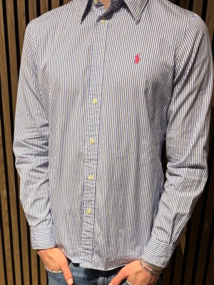 Randig skjorta från Polo Ralph Lauren - Snygg blåvit randig skjorta från Polo Ralph Lauren med klassisk krage och knappar framtill. Står storlek 10 men det är samma som S. Custom fit-modell med lång ärm och ikonisk rosa logga broderad på bröstet. Perfekt till jeans eller chinos för en clean och stilren look.
