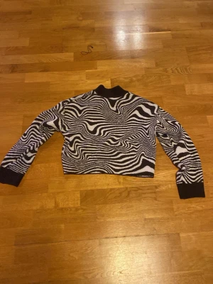 Zebramönstrad stickad tröja - Säljer en cool zebramönstrad stickad tröja i svart och vitt. Tröjan har hög krage, långa ärmar och ribbade muddar vid ärmslut och nederkant. Perfekt för dig som vill sticka ut med ett unikt mönster!