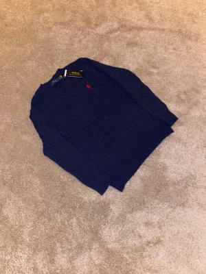 Mörkblå stickad tröja Polo Ralph Lauren - Klassisk mörkblå stickad tröja från Polo Ralph Lauren med rund halsringning och det ikoniska röda broderade logot på bröstet. Tröjan har kabelstickat mönster och långa ärmar, perfekt för en stilren och tidlös look.