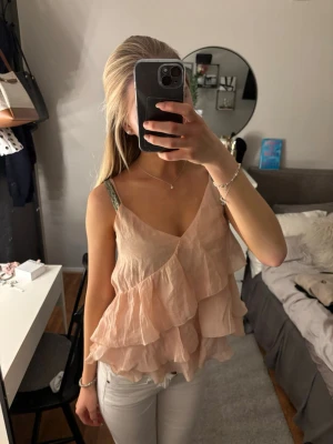 Ljusrosa volangig linnetopp - Säljer en superfin ljusrosa linnetopp med flera lager volanger och tunna axelband. Toppen har en v-ringad halsringning och ett luftigt, flowy material som känns perfekt för varmare dagar. Passar dig som gillar en romantisk och söt stil.