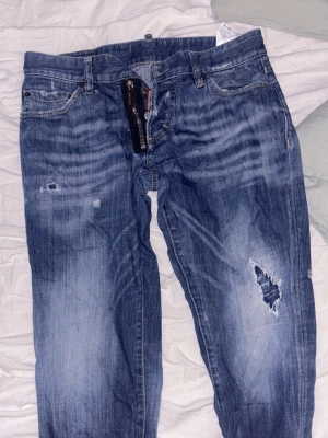 Blå slitna jeans med hål - Säljer ett par blå jeans med slitningar och hål på ena benet. Jeansen har en klassisk femficksmodell, dragkedja och knapp i midjan. Materialet är denim och de har en tvättad look med ljusare partier och distressed detaljer.