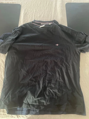 Svart t-shirt från Gant - Klassisk svart t-shirt från Gant med liten broderad logga på bröstet. T-shirten har rund halsringning och korta ärmar. Perfekt basplagg till garderoben och enkel att matcha med allt.