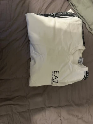 Vit EA7 sweatshirt med logga - Snygg vit sweatshirt från EA7 Emporio Armani med svarta logoband längs ärmarna och tryckt logga på bröstet. Tröjan har rund halsringning och långa ärmar. Perfekt för en clean och sportig look.