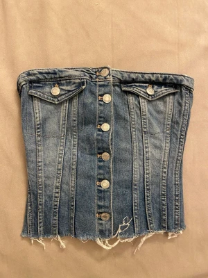 Populär jeans korsettopp från Zara - Säljer en snygg jeans korsettopp från Zara med knappar framtill och två dekorativa bröstfickor. Toppen är axelbandslös och har rå kant nertill för en edgy look. Perfekt till sommaren för en snygg sommar outfit!