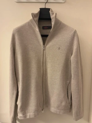 Beige ziptröja från Morris Stockholm - Snygg beige ziptröja från Morris Stockholm med hög krage och diskret logga på bröstet. Tröjan har hel dragkedja, två fickor och är tillverkad i mjukt stickat material. Perfekt för lager-på-lager och stilren look. 