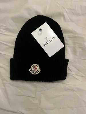 Svart Moncler mössa  - Snygg svart ribbstickad mössa från Moncler med uppvikt kant och klassisk Moncler-logga framtill. Tillverkad i mjukt material som håller dig varm och ger en clean look. Perfekt accessoar för kalla dagar. Man får med original förpackning kom privat för pris och andra frågor 👍🏼