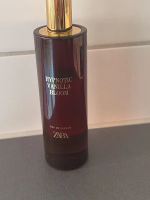 Zara Hypnotic Vanilla Bloom parfym - En elegant parfymflaska från Zara med doften Hypnotic Vanilla Bloom. Flaskan är mörkbrun med gulddetaljer och har en rund, stilren form. Texten på flaskan är i guld och locket är också guldfärgat, vilket ger en lyxig känsla.