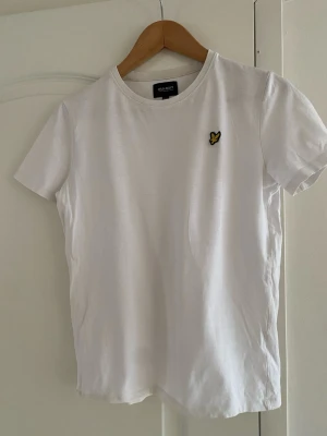 Vit t-shirt från Lyle & Scott - Klassisk vit t-shirt från Lyle & Scott med rund hals och korta ärmar. På bröstet sitter den ikoniska gula örn-loggan som ger en stilren touch. Perfekt basplagg i mjuk bomull som funkar till allt.