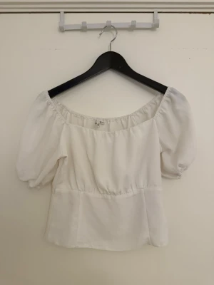 Vit offshoulder blus med puffärm - Söt vit blus med offshoulder-design och puffiga korta ärmar. Toppen har en lätt rynkad midja och är tillverkad i ett mjukt tyg som ger en romantisk vibe. Perfekt att styla med jeans eller kjol för en trendig look.