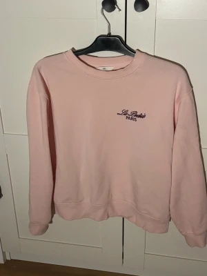 Ljusrosa sweatshirt från H&M - Ljusrosa sweatshirt från H&M med broderad text 'Le Paris PARIS' i svart på bröstet. Tröjan har rund halsringning, långa ärmar och ribbade muddar vid ärmslut och nederkant. Perfekt för en avslappnad och trendig look.