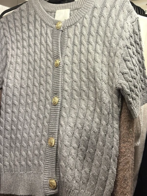 Grå kabelstickad kofta från H&M - Kortärmad grå kabelstickad kofta från H&M med rund halsringning och dekorativa guldfärgade knappar framtill. Tröjan har ribbade muddar och ett klassiskt kabelmönster som ger en trendig och stilren look. Perfekt att styla över en topp eller klänning.