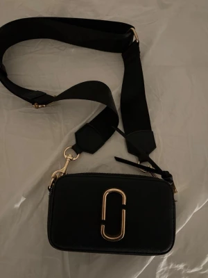 Svart axelväska Marc Jacobs - Snygg svart axelväska från Marc Jacobs med bred justerbar axelrem och guldiga metalldetaljer. Väskan har en rektangulär form, dubbla dragkedjor och den ikoniska loggan framtill. Tillverkad i läder med detaljer i metall.