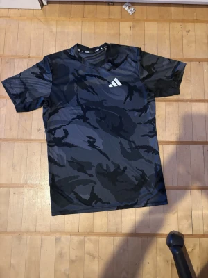 Svart camo t-shirt från Adidas - Snygg svart och mörkgrå t-shirt från Adidas med camomönster och vitt Adidas-logga på bröstet. Tillverkad i ett lätt och snabbtorkande material, perfekt för träning eller sportaktiviteter. Kortärmad och med rund hals.