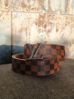 Brun/rutigt bälte med metallspänne - Säljer ett brunt bälte med klassiskt rutigt mönster och ett stort märke i metall. Bältet har en lyxig känsla och passar perfekt till jeans eller kostymbyxor. Materialet är skinn och designen är riktigt ikonisk.