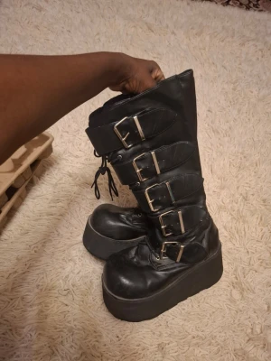 Svarta platåstövletter med spännen - Tuffa svarta stövletter i skinnimitation med kraftig platåsula och fyra stora metallspännen på sidan. Snörning framtill för extra edge och justerbar passform. Perfekta för dig som gillar alternativ stil och vill sticka ut.