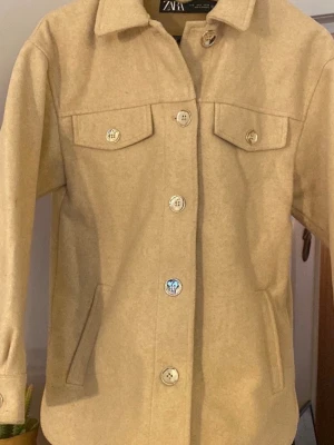Beige jacka/skjorta från ZARA - Stilren beige jacka/skjorta från ZARA med klassisk krage, stora knappar och två bröstfickor med lock. Tillverkad i ett mjukt material som ger en avslappnad men ändå snygg look. Perfekt att bära över en hoodie eller t-shirt för extra lager.