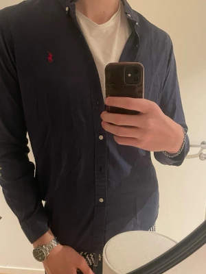 Mörkblå skjorta från Polo Ralph Lauren - Säljer en mörkblå skjorta från Polo Ralph Lauren i slim fit. Skjortan har långa ärmar, klassisk krage och knappar framtill. Ikoniska röda loggan broderad på bröstet. Perfekt för dig som gillar stilrena och tidlösa plagg. Saknar en knapp!