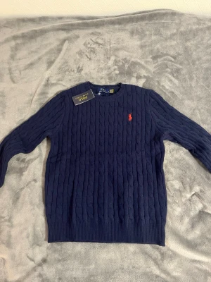 Mörkblå kabelstickad tröja Polo Ralph Lauren - Kabelstickad mörkblå tröja från Polo Ralph Lauren med klassisk röd broderad logga på bröstet. Tröjan har rund halsringning, ribbade muddar och är tillverkad i mjukt material. Perfekt för en stilren och avslappnad look.