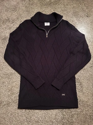 Marinblå stickad tröja med dragkedja SAMS - Marinblå stickad tröja från SAMS med halv dragkedja och hög krage. Snyggt mönsterstickad yta i geometriskt zigzag-mönster. Ribbstickade muddar och nederkant. Perfekt för lager-på-lager och stilren look.