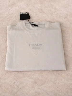 Vit t-shirt från Prada med logga - Vit t-shirt från Prada med klassisk rund hals och korta ärmar. Framsidan har en snygg, broderad Prada Milano-logga i triangelform. Materialet känns mjukt och exklusivt, perfekt för dig som gillar stilrena designer och lyxiga märken. Äkta med prislapp. Aldrig använd 