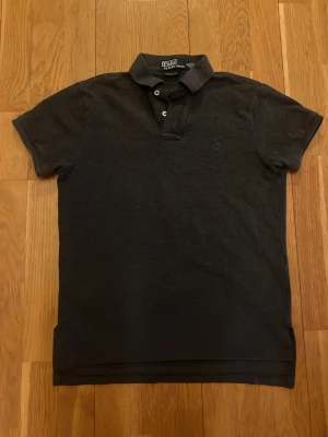 Grå pikétröja från Ralph Lauren - Klassisk grå pikétröja från Ralph Lauren med broderad grön logga på bröstet. Tröjan har korta ärmar, krage och knäppning med två knappar. Perfekt för en stilren och laidback look på sommaren eller våren.