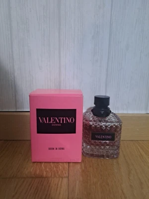 Valentino Donna Born In Roma parfym - Valentino Donna Born In Roma parfym med en lyxig, rosa glasflaska med nitar och svart lock. Förpackningen är starkt rosa med svart etikett och vit text. Flaskan har en elegant och modern design som känns exklusiv och trendig.