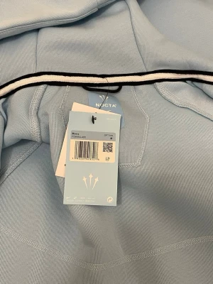 Ljusblå NOCTA hoodie från Nike - Snygg ljusblå hoodie från Nike NOCTA med dragkedja framtill, två fickor med dragkedja och broderad Nike-logga på bröstet. Hoodien har huva och diskret NOCTA-tryck på ärmen. Perfekt för en stilren och sportig look.