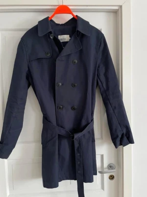 Mörkblå trenchcoat med bälte pier ome - Stilren mörkblå trenchcoat med dubbelknäppning och klassisk krage. Jackan har axelklaffar, bälte i midjan och knappar vid ärmsluten. Tillverkad i ett lätt tyg som passar perfekt till vår och höst. Snygg och tidlös modell som funkar till många olika looks.