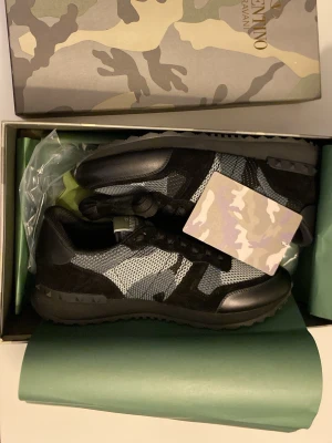 Valentino Garavani rockrunners  - Snygga svarta sneakers från Valentino Garavani med mesh och mockadetaljer. Skorna har kamouflagemönster i grått och svart, samt svarta snören och gummisula med nitar. Perfekta för dig som vill sticka ut med stilrena och exklusiva sneakers. Pris kan diskuteras😊