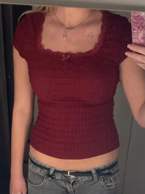 Vinröd croppad topp med rosett - Säljer en vinröd croppad topp med korta puffärmar och söt liten rosett framtill. Toppen har smockad struktur och en fin, vågig kant vid urringningen. Perfekt för dig som gillar en feminin och trendig stil.