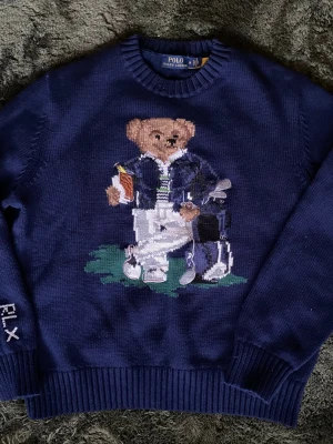 Mörkblå Polo Ralph Lauren stickad tröja - Mörkblå stickad tröja från Polo Ralph Lauren med ikonisk björn i golfoutfit broderad på bröstet. Tröjan har rund hals, ribbade muddar och RLX-detalj på ärmen. Perfekt för dig som gillar preppy stil och unika detaljer.