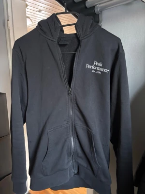 Svart hoodie från Peak Performance - Jag säljer min snygga svarta zip-hoodie från Peak Performance med vit logga och text på bröstet. Klassisk modell med dragkedja, två fickor framtill och stor huva. Tillverkad i mjukt bomullsmaterial. Hör av er vid frågor! 