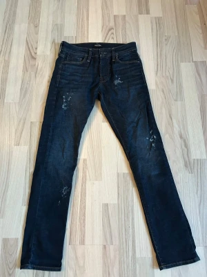 Mörkblå slim fit jeans med slitningar - Snygga mörkblå slim fit jeans från Jack & Jones med diskreta slitningar framtill och bakfickor. Klassisk femficksmodell med knappgylf och läderpatch bak i midjan. Tillverkade i bomull med lite stretch för skön passform.