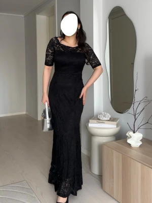 Svart lång spetsklänning - Elegant svart långklänning med figurnära passform och halvärmade ärmar i spets. Klänningen har rund halsringning och vackra spetsdetaljer över axlar och ärmar. Perfekt för en stilren look med en touch av romantik.