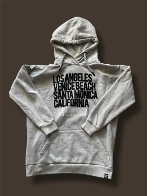 Grå hoodie från Superdry - Snygg grå hoodie från Superdry med svart texttryck på bröstet där det står Los Angeles, Venice Beach, Santa Monica, California. Klassisk känguruficka och justerbar huva med snören. Perfekt för en avslappnad streetwear-look. 