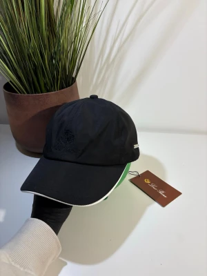 Loro Piana keps - Stilren svart keps från Loro Piana med diskret broderad logga framtill och liten metallbricka på sidan. Kepsen har böjd skärm med vit kant och är tillverkad i ett lätt, troligtvis bomulls- eller nylonmaterial. Perfekt accessoar för en clean och modern look.