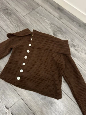 Brun stickad kofta med sned knäppning - Snygg brun stickad kofta med lång ärm och unik sned knäppning framtill med vita knappar. Koftan har en bred krage och är virkad i ett grovt mönster. Perfekt för lager-på-lager och chill stil. I storlek M
