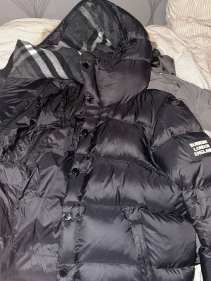 Svart pufferjacka från Burberry - Snygg svart pufferjacka från Burberry med huva och klassiskt rutigt innerfoder. Jackan har dragkedja och knappar framtill samt praktiska fickor med dragkedja. Märkespatch på ärmen och quiltad design för extra värme. Den är knappt använd har bara testat den.