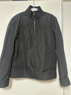 Michael kors windbreaker - Stilren svart vindjacka med ståkrage och hel dragkedja framtill. Två sidofickor och justerbara ärmslut. Jackan har en minimalistisk design utan synligt märke och är tillverkad i ett slätt, vindtätt material. Perfekt för blåsiga dagar.