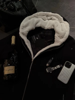 Moose Knuckles Classic Bunny hoodie / jacka - Snygg svart stilren ”Classic Bunny” hoodie / jacka från Moose Knuckles med stor huva fodrad i vit päls. Kappan har dragkedja framtill och den ikoniska loggan på vänster ärmen. Perfekt för kalla dagar när du vill hålla stilen och värmen. Passar mig som brukar använda M.